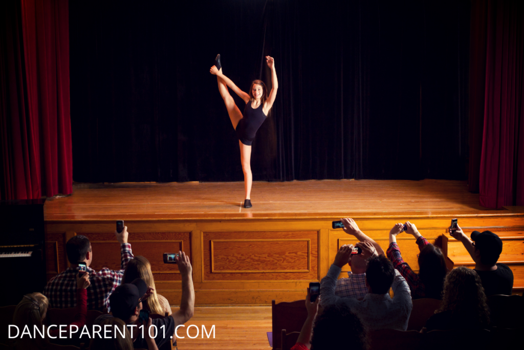Girl on stage holding one leg high in the air and people in the audience using phones to film her - Dance Recital