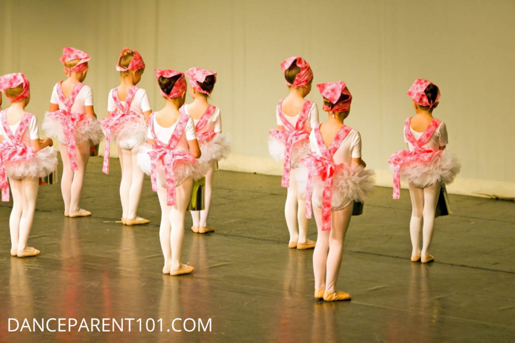 We see the backs of a group of young dancers in costume on stage about to perform in their dance recital