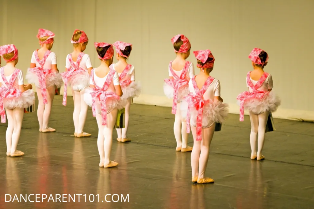 We see the backs of a group of young dancers in costume on stage about to perform in their dance recital