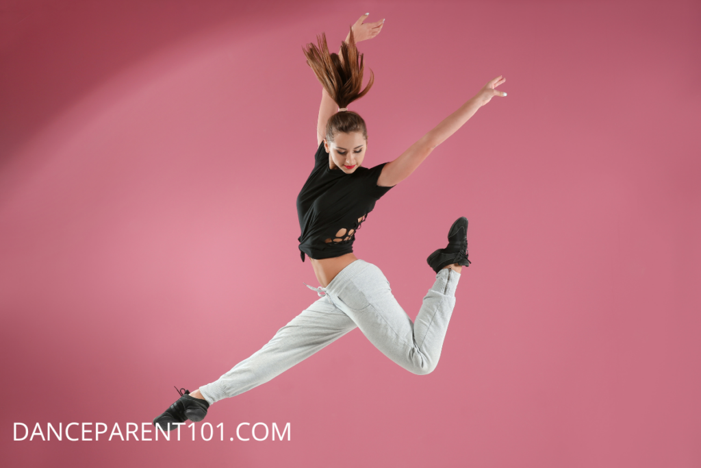 Female dancer jumping in front of a pink background wearing grey sweats, a black shirt and black dance sneakers