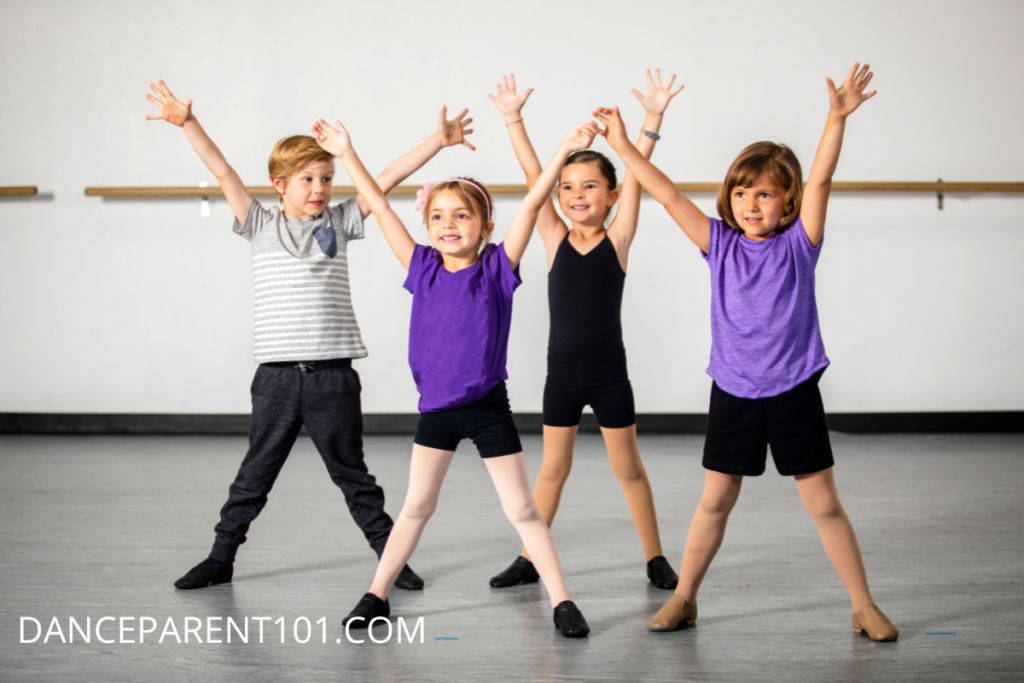 A group of 4 children wearing purple, gray, and black outfits with jazz shoes stand facing the camera with arms outstretched