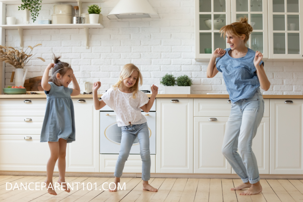 A mom and two young girls are dancing in a white and tan kitchen. They are wearing various shades of denim