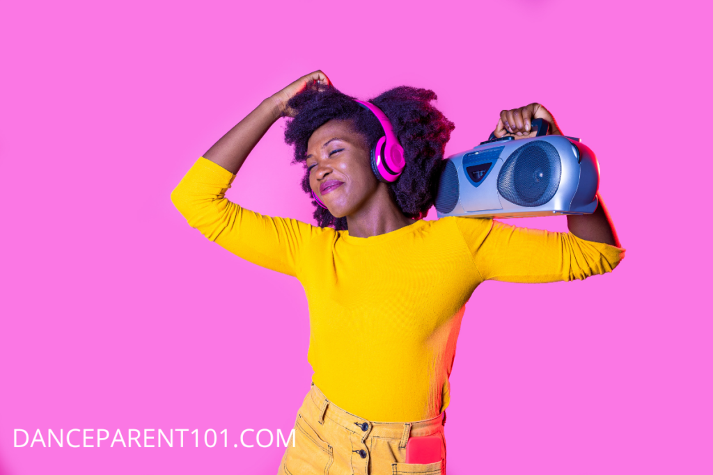 A woman wearing a bright yellow shirt and pants and pink headphones holds a blue boombox on her shoulder against a bright pink background