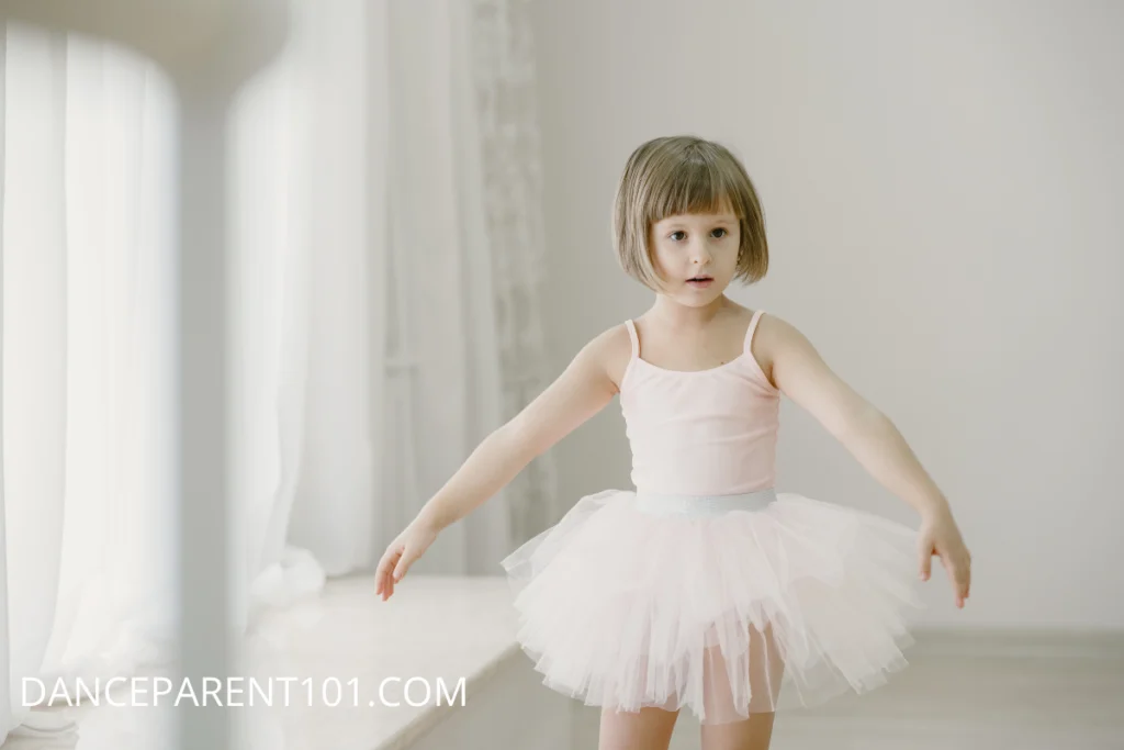 A little girl with short blonde hair stands in a dance studio. She is wearing a light pink leotard and tutu and holding her arms out by her side.