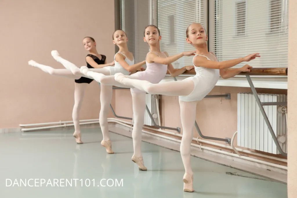 A line of young ballerinas standing en attitude at the barre. They are wearing white and black leotards and pink tights.