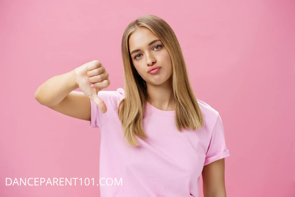 A blonde teen girl wearing a pink shirt against a pink background gives a thumbs down sign