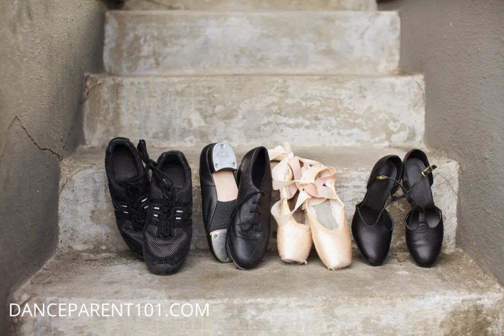 Image of 4 pairs of dance shoes sitting on stairs, from left, black hip hop sneakers, black oxford tap shoes, pink pointe shoes, and black character shoes