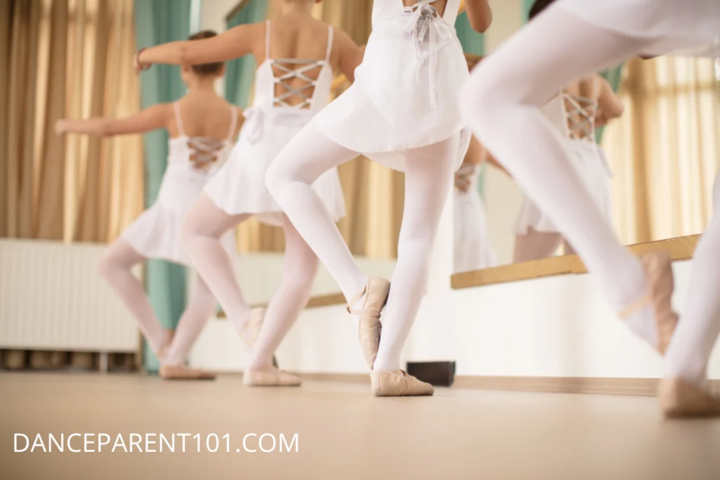 Image of young dancers facing away from the camera. They wear white leotards and skirts, tights and ballet shoes and are standing at the barre in sur la coup de pied position.